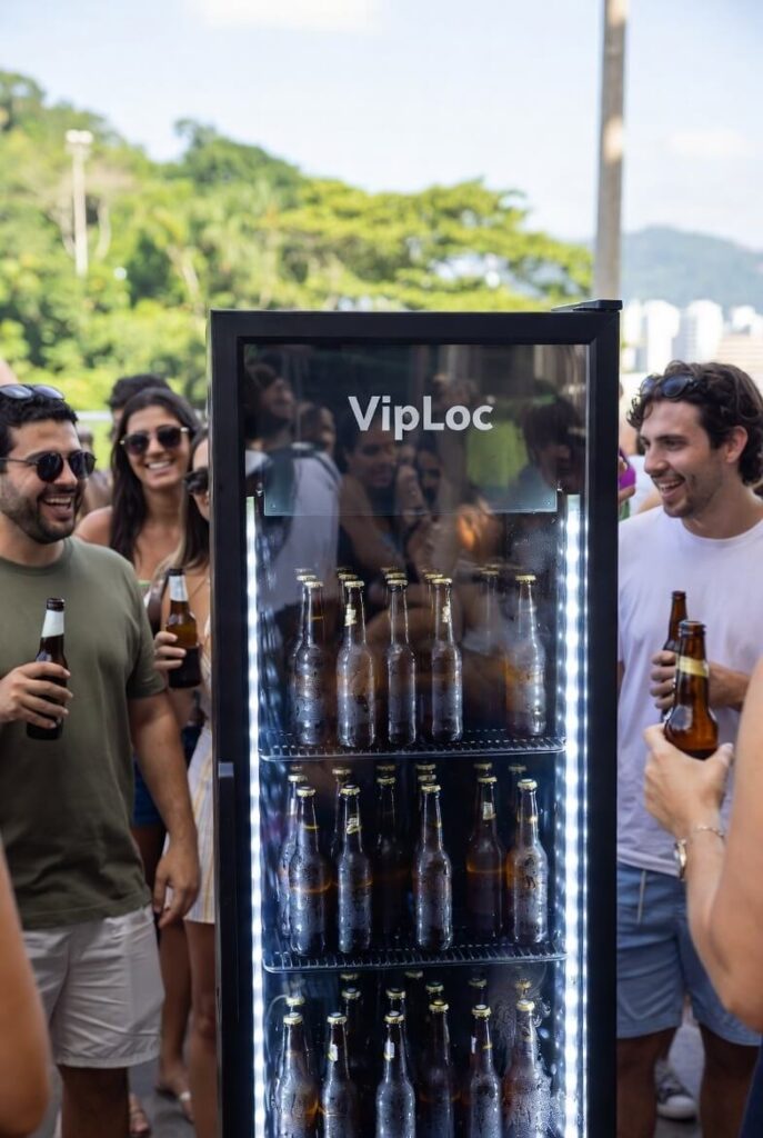Grupo de pessoas comemorando ao redor de uma geladeira de cerveja, com destaque para a marca VipLoc, em um evento ao ar livre sob céu claro.